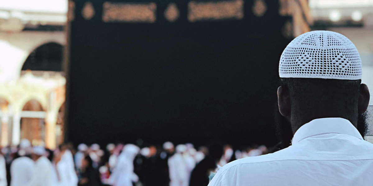 Un homme de dos portant une chéchia blanche se tient face à la Kaaba à Masjid al-Haram, illustrant la réalisation d'une Omra par procuration.