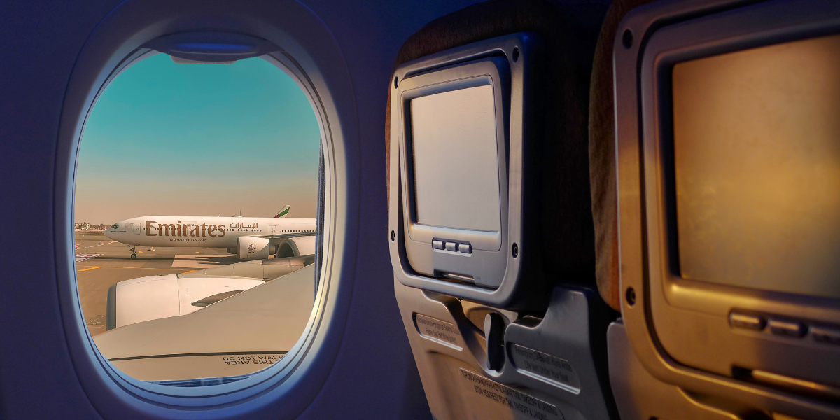 Vue depuis la cabine d'un avion Emirates montrant les écrans individuels et, par le hublot, un autre appareil de la flotte sur le tarmac, illustrant l'expérience de vol premium pour les pèlerins.