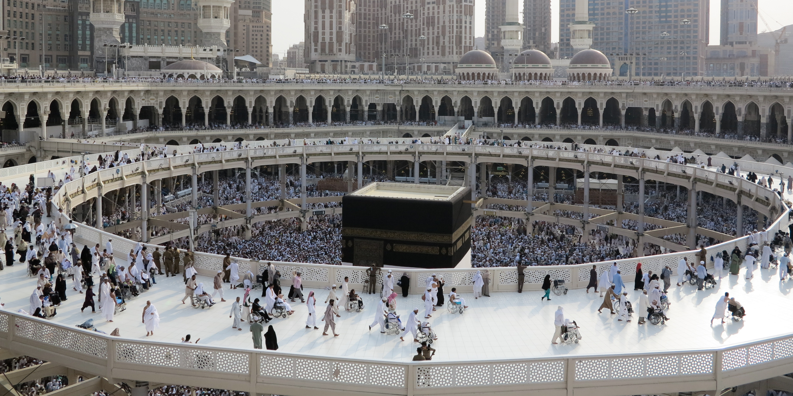Vue en plongée de la Kaaba à La Mecque, entourée par les passerelles circulaires de la Mosquée al-Haram où cheminent des pèlerins, illustrant l'équilibre parfait entre l'autonomie du pèlerin et l'assistance d'un guide sur place pour optimiser son temps et son budget.