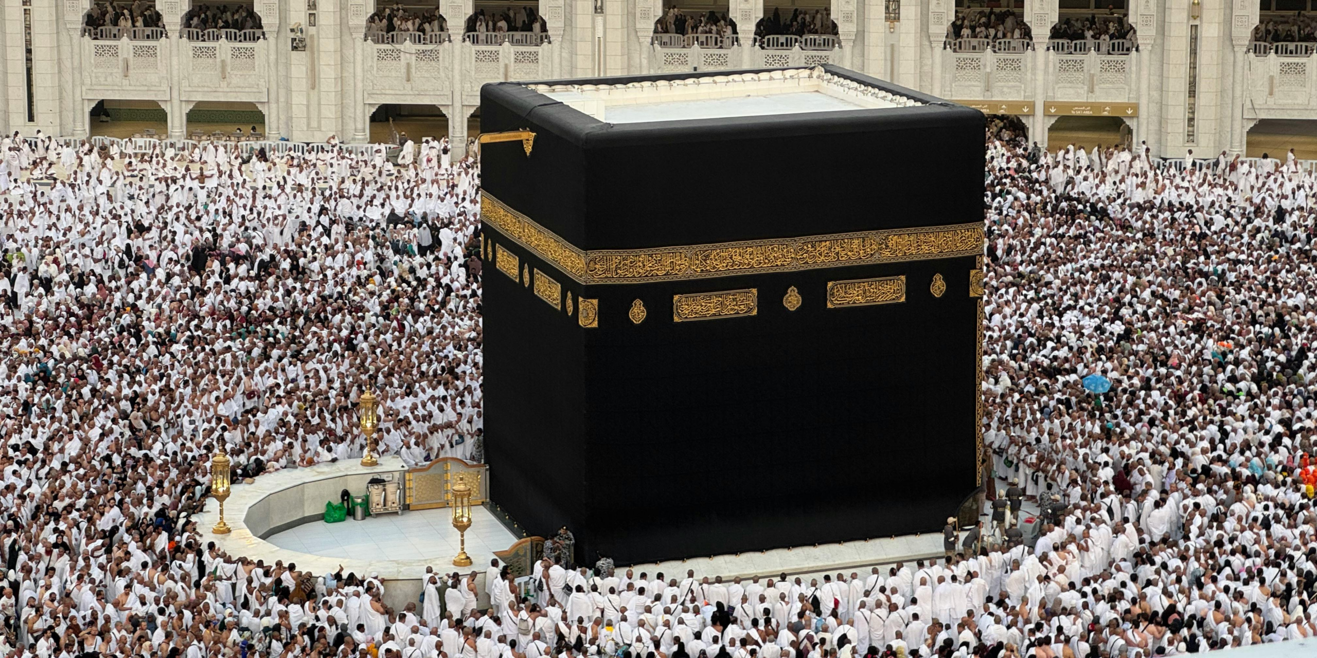 Vue rapprochée en plongée de la Kaaba drapée de sa Kiswa noire et dorée, entourée d'une foule dense de pèlerins en habit d’ihram à La Mecque, illustrant parfaitement les défis logistiques et la gestion de la foule pour un pèlerin effectuant son Omra en solo.
