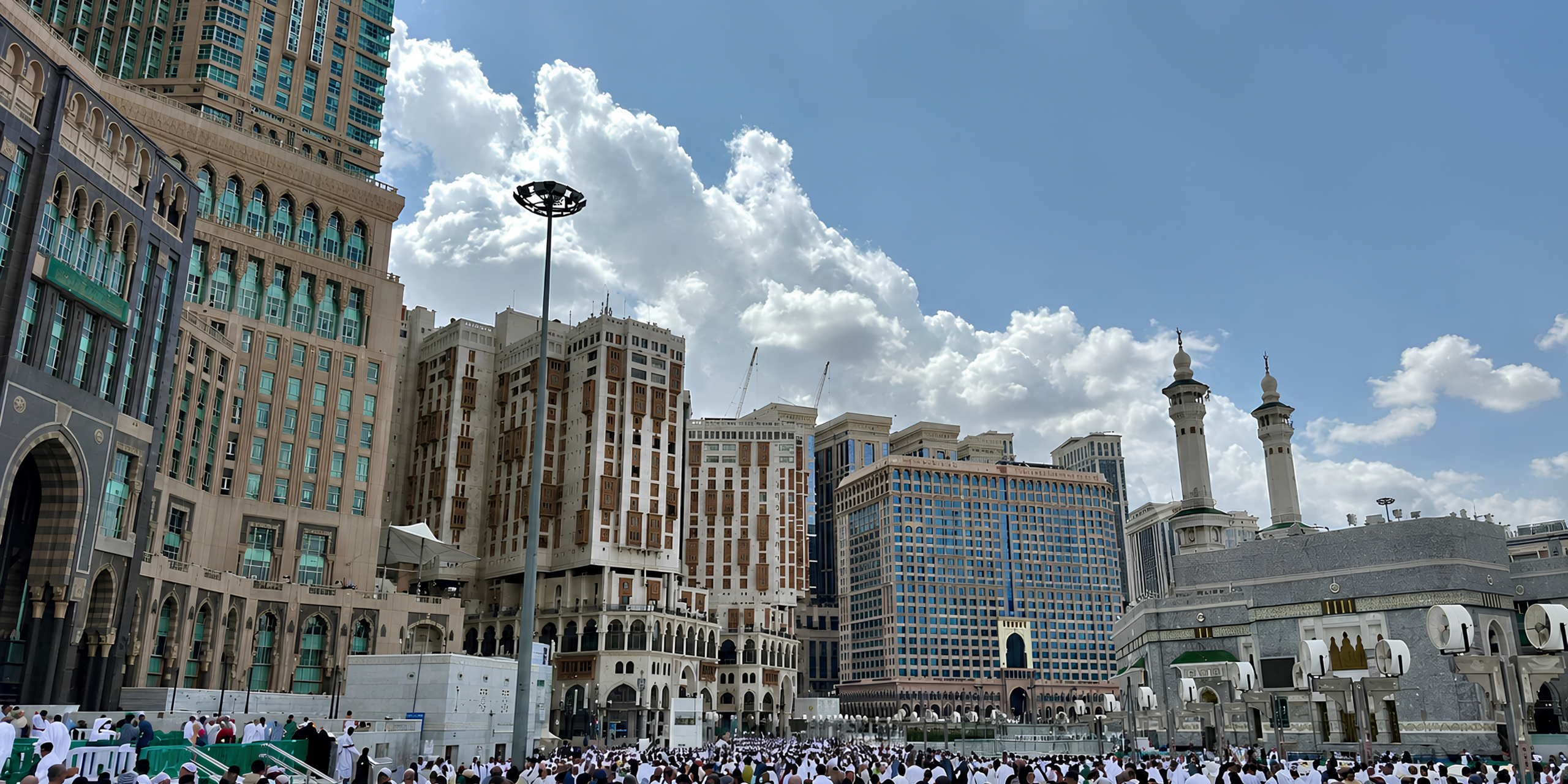 Vue extérieure depuis l'esplanade de la Grande Mosquée de La Mecque montrant les grands hôtels et les bâtiments environnants sous un ciel nuageux, illustrant la préparation logistique, les documents indispensables et les conseils pratiques pour un premier voyage en Omra.