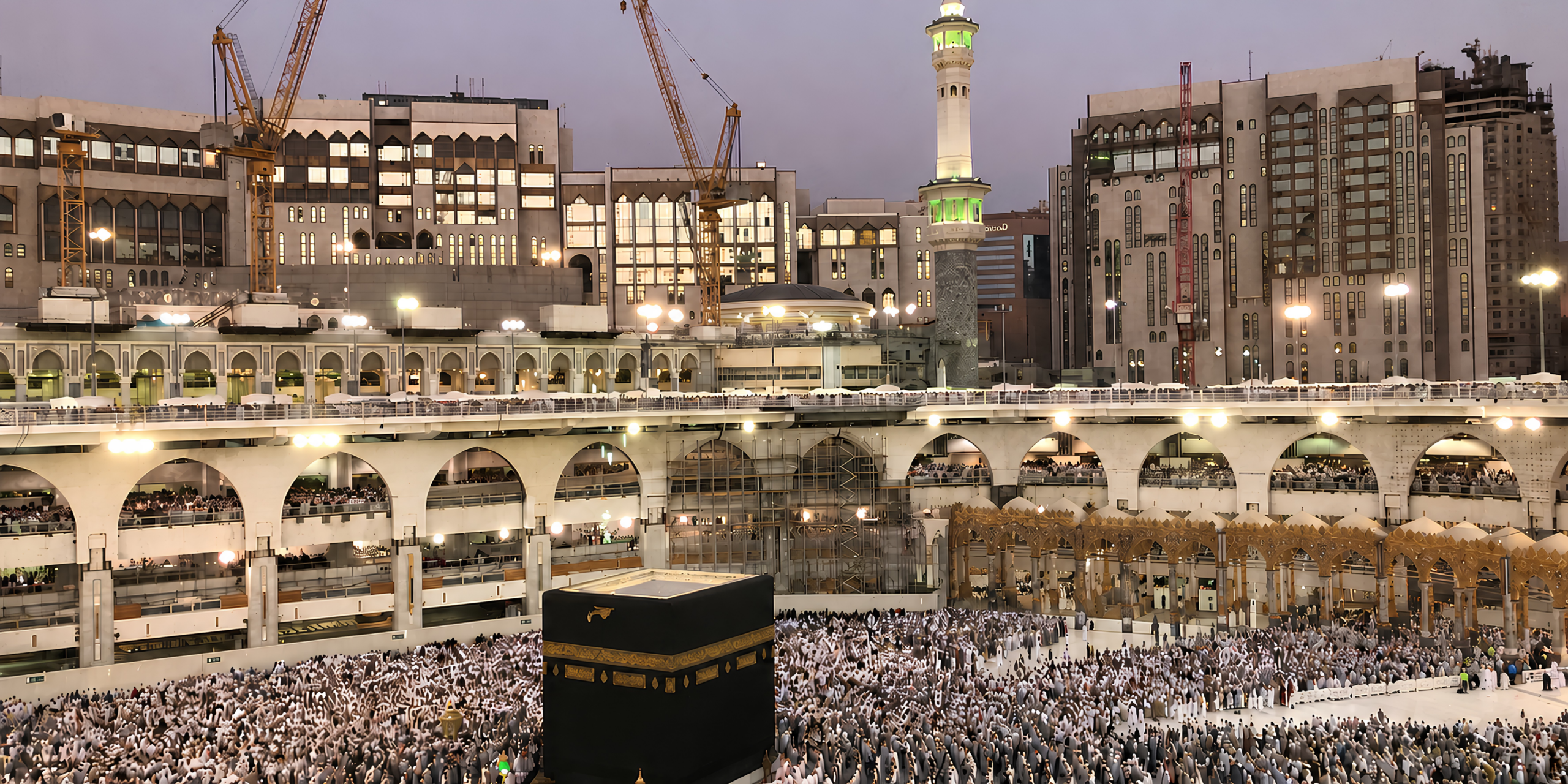 Vue sur la Kaaba et la cour de la Grande Mosquée de La Mecque s'illuminant au crépuscule avec les pèlerins en arrière-plan, illustrant les réponses aux questions logistiques, de visa et d'organisation des débutants.