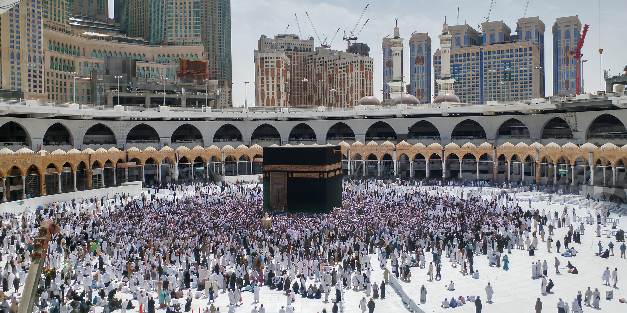 Vue panoramique en journée de l'esplanade de la Grande Mosquée de La Mecque, montrant les pèlerins vêtus d'Ihram rassemblés autour de la Kaaba sous un grand soleil, illustrant la définition et l'importance spirituelle des premières étapes de la Omra pour les débutants.