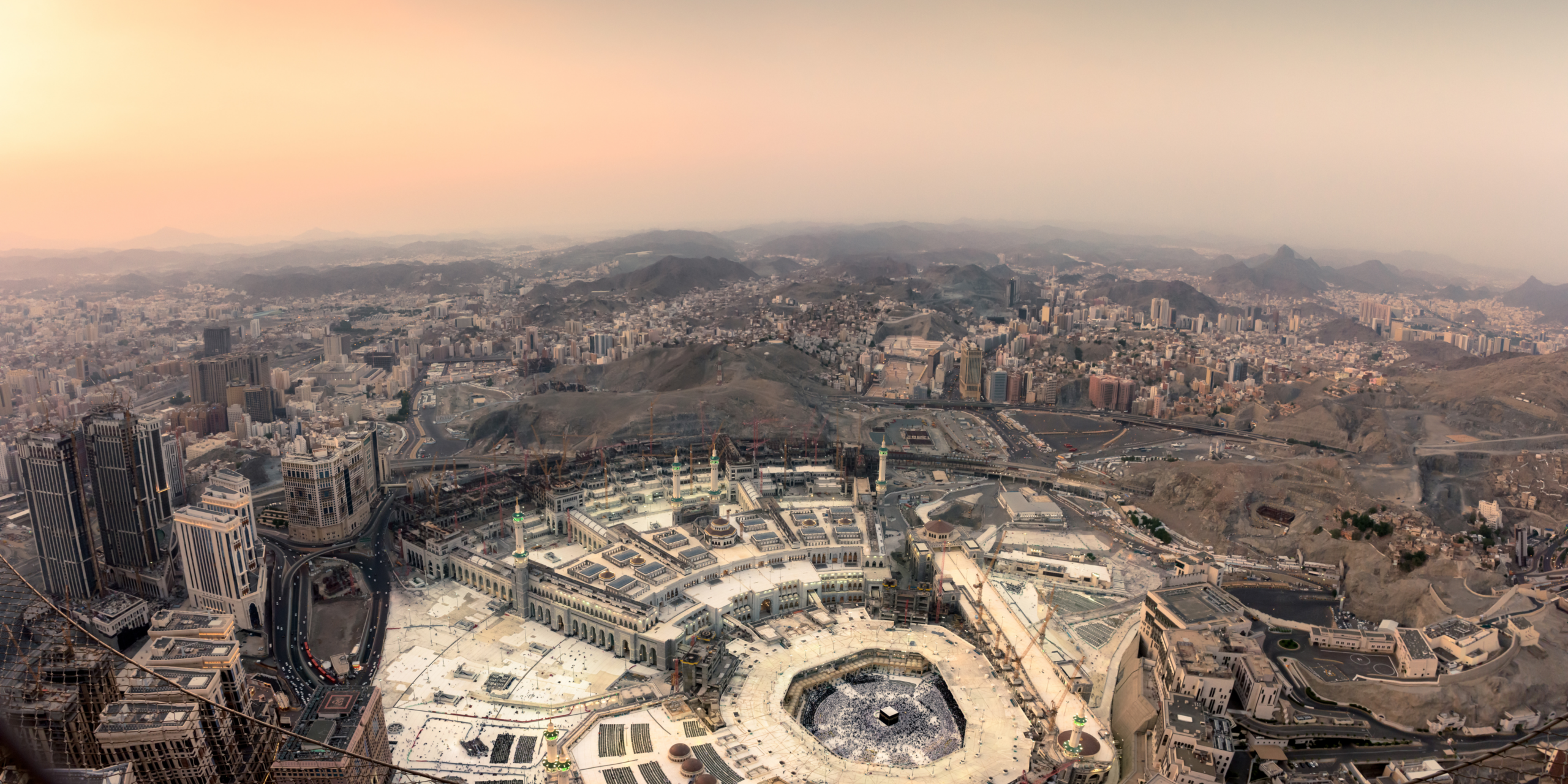 Vue aérienne panoramique de la Mosquée Al-Haram et de la Kaaba à la Mecque au coucher du soleil, entourées par les gratte-ciels, la ville et les collines rocheuses en arrière-plan sous un ciel dégradé de jaune et de beige, illustrant l'expertise et le confort d'un service de guide Omra premium à La Mecque.