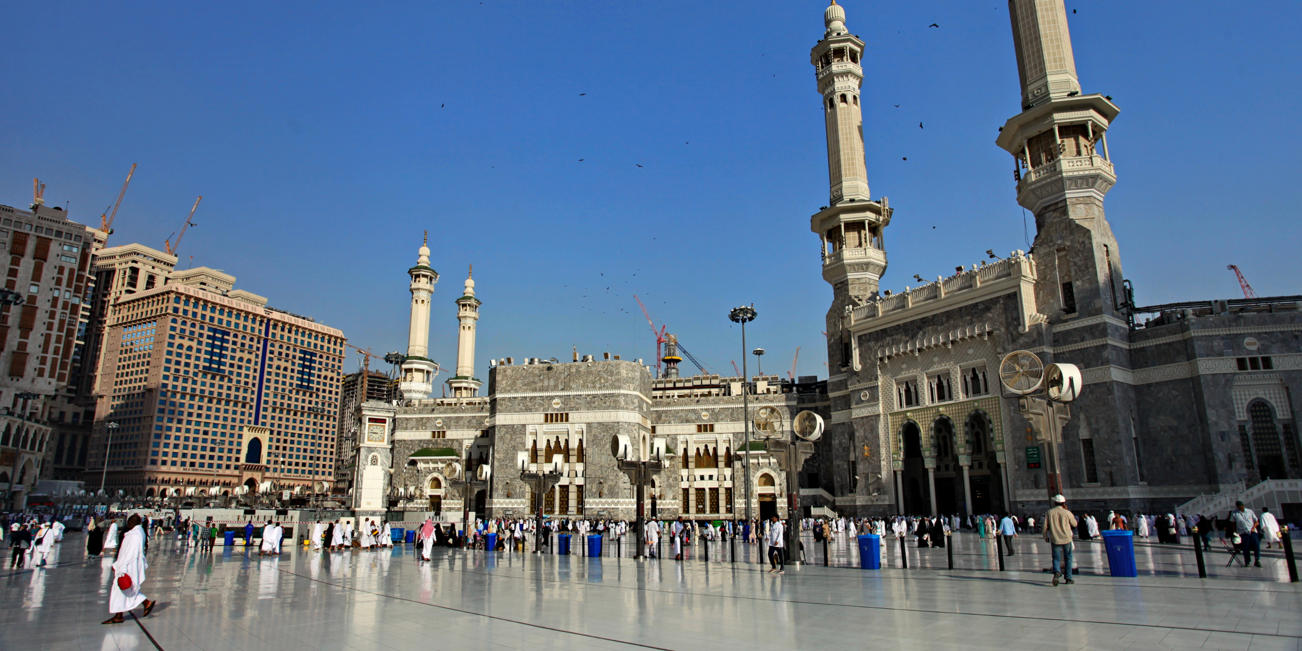 Pèlerins marchant sur la grande esplanade en marbre de Masjid al-Haram devant des hôtels de standing, illustrant la logistique et l'hébergement clé en main des services Direct Mecca.