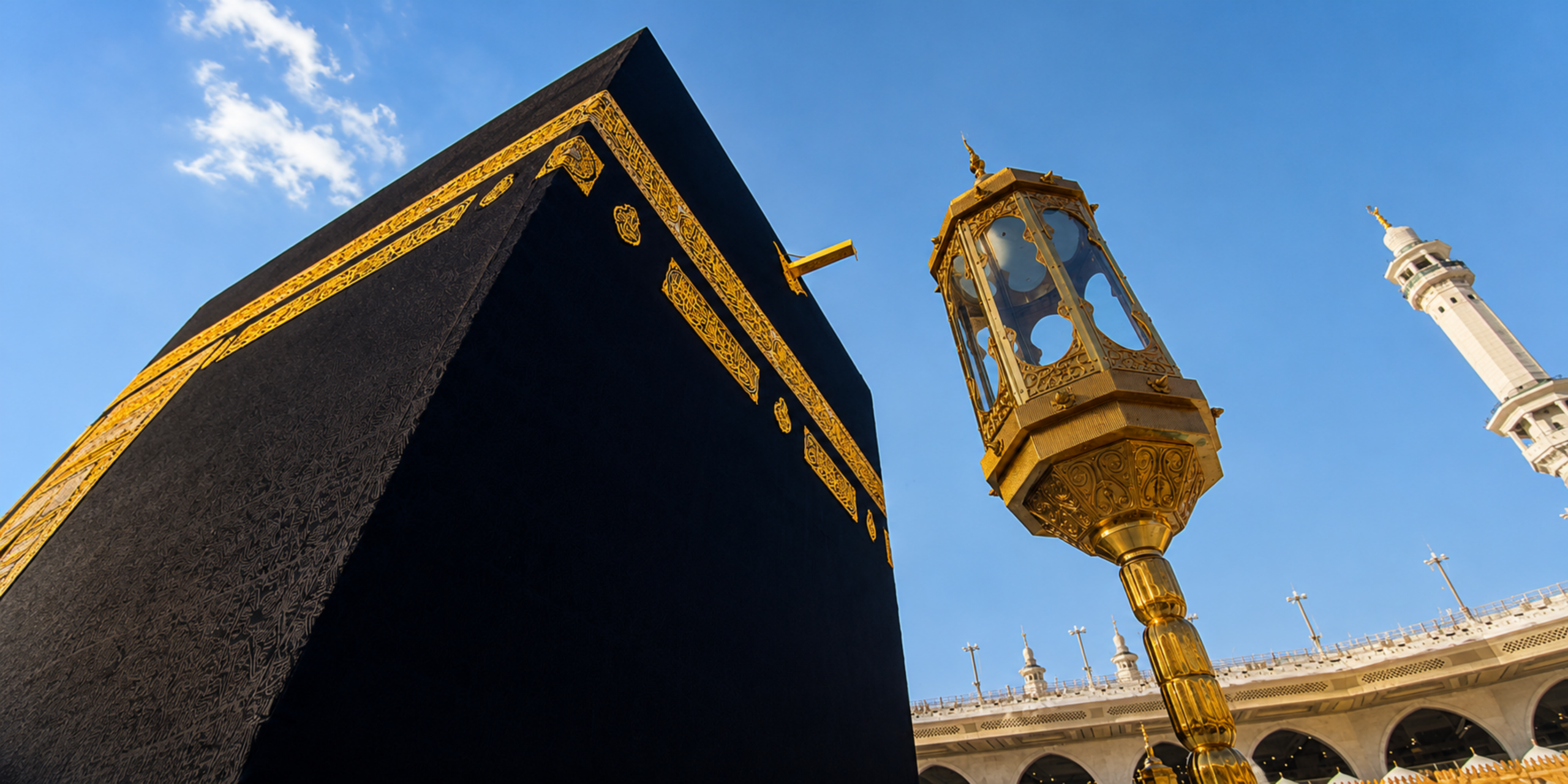 Une vue en contre-plongée de la Kaaba drapée de sa Kiswa noire aux broderies dorées à La Mecque, à côté d'un grand lampadaire doré de style traditionnel sous un ciel bleu lumineux, illustrant la planification d'un séjour de courte durée avec un Guide Omra express.