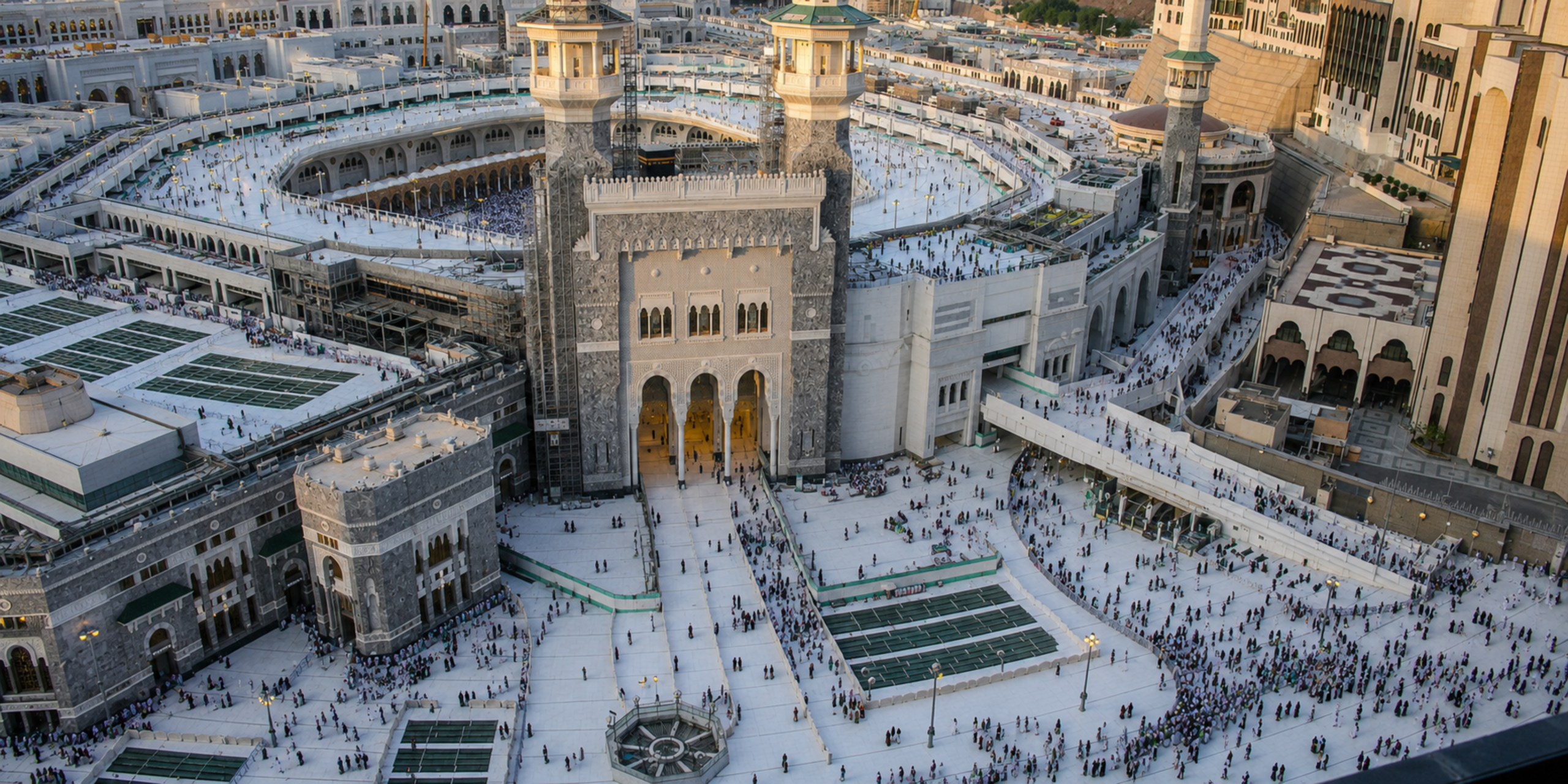 Une vue aérienne plongeante sur l'une des entrées monumentales de la Grande Mosquée de La Mecque (Al-Masjid al-Haram), montrant l'architecture blanche et grise du complexe, les esplanades extérieures et les pèlerins qui circulent, illustrant la complexité de la gestion logistique entre un voyage en solo et un séjour encadré par un Guide Omra express.
