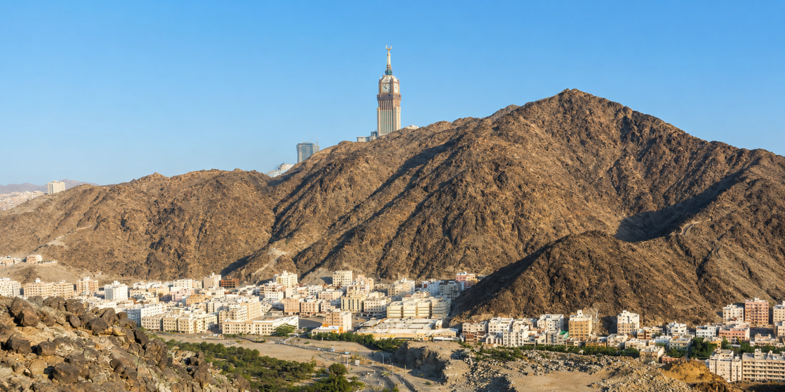 Un paysage panoramique de La Mecque montrant un quartier résidentiel niché au pied d'une montagne rocheuse aride, surmontée au loin par la Tour de l'Horloge (Makkah Clock Royal Tower) sous un ciel bleu clair, illustrant la prise en charge logistique et l'hébergement avec les services Direct Mecca.