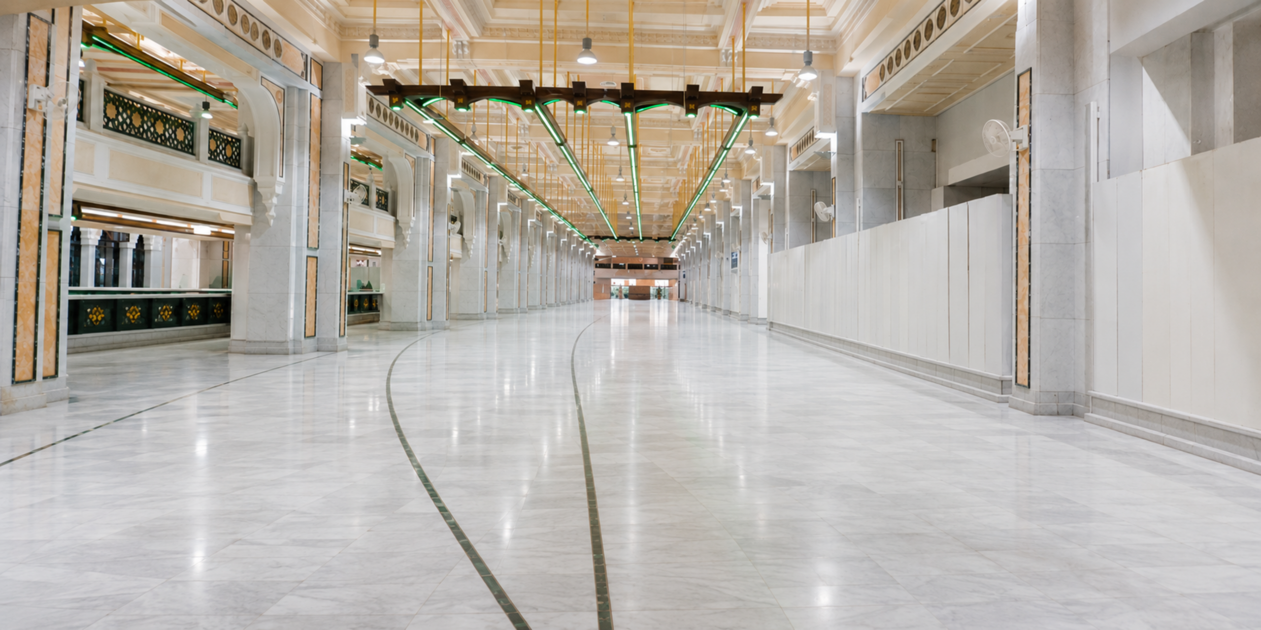 Une vue en perspective de la galerie intérieure en marbre blanc de Safa et Marwah (le Mas'a) au sein de la Grande Mosquée de La Mecque, montrant un couloir spacieux et vide flanqué de piliers monumentaux, avec des structures de signalisation vertes suspendues au plafond et des lignes de guidage tracées au sol, illustrant la préparation logistique et spirituelle avant le départ en omra avec un guide.