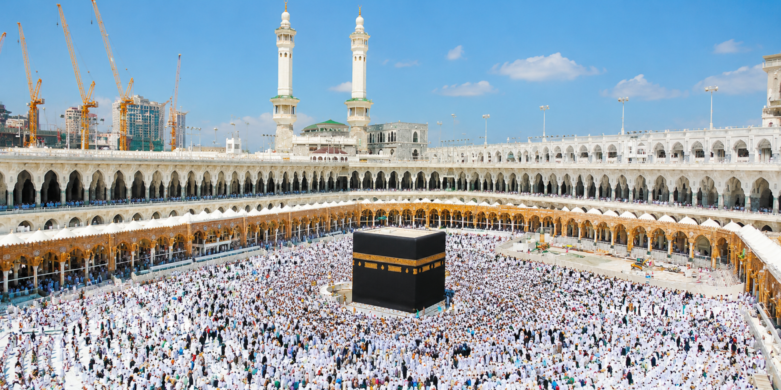 Une vue panoramique en plongée de la cour bondée de la Grande Mosquée de La Mecque (Al-Masjid al-Haram) avec une multitude de fidèles entourant la Kaaba, encadrée par les galeries à arcades et deux grands minarets blancs sous un ciel azur, illustrant la recherche et l'anticipation d'un Guide Omra express dans cet espace d'affluence.