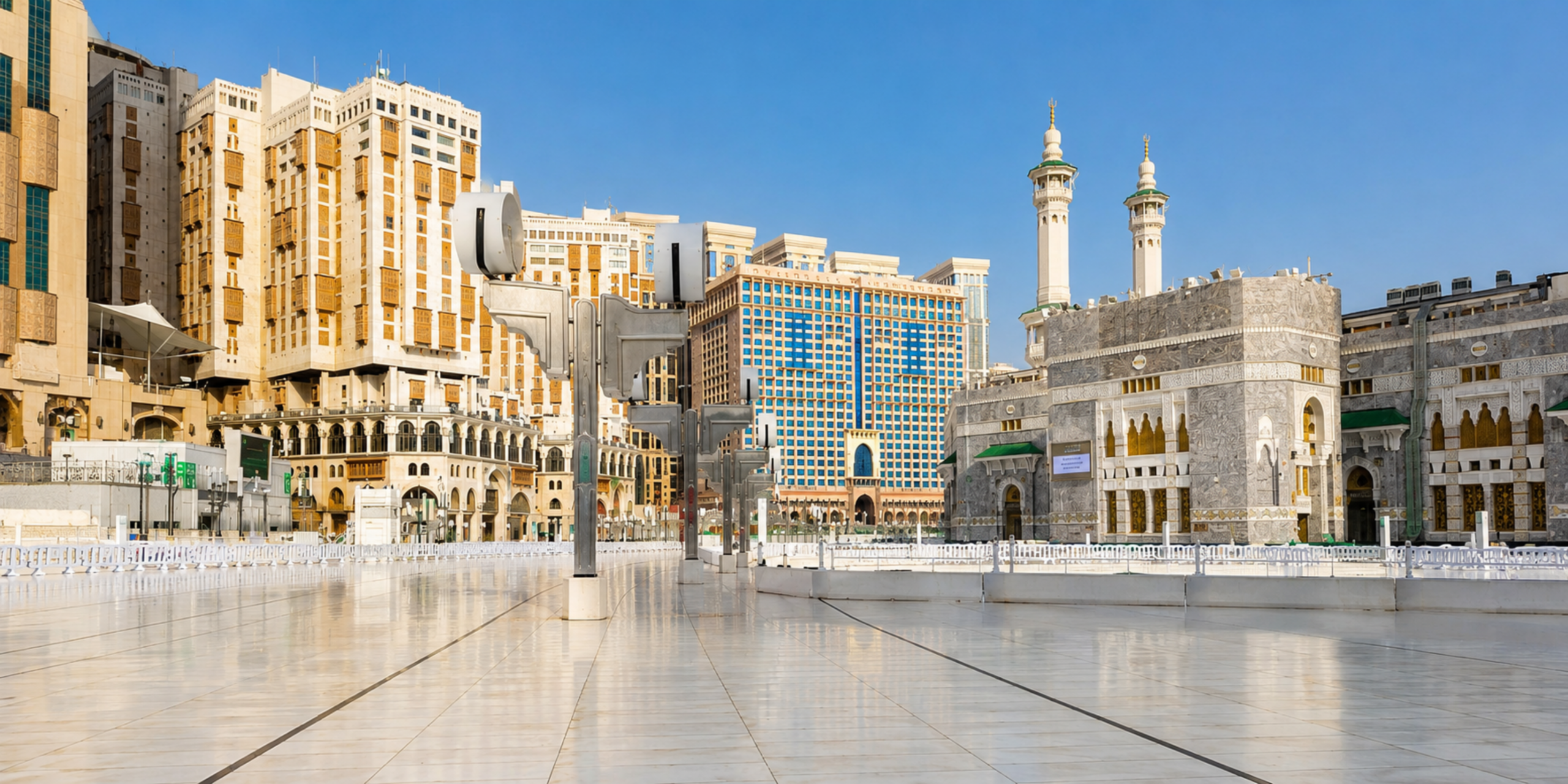 Une vue de plain-pied sur l'esplanade extérieure dallée de marbre blanc de la Grande Mosquée de La Mecque, bordée de grands immeubles et d'hôtels de standing sous un ciel bleu, illustrant les options d'hébergement sur mesure et de logistique personnalisée à proximité immédiate du Haram.
