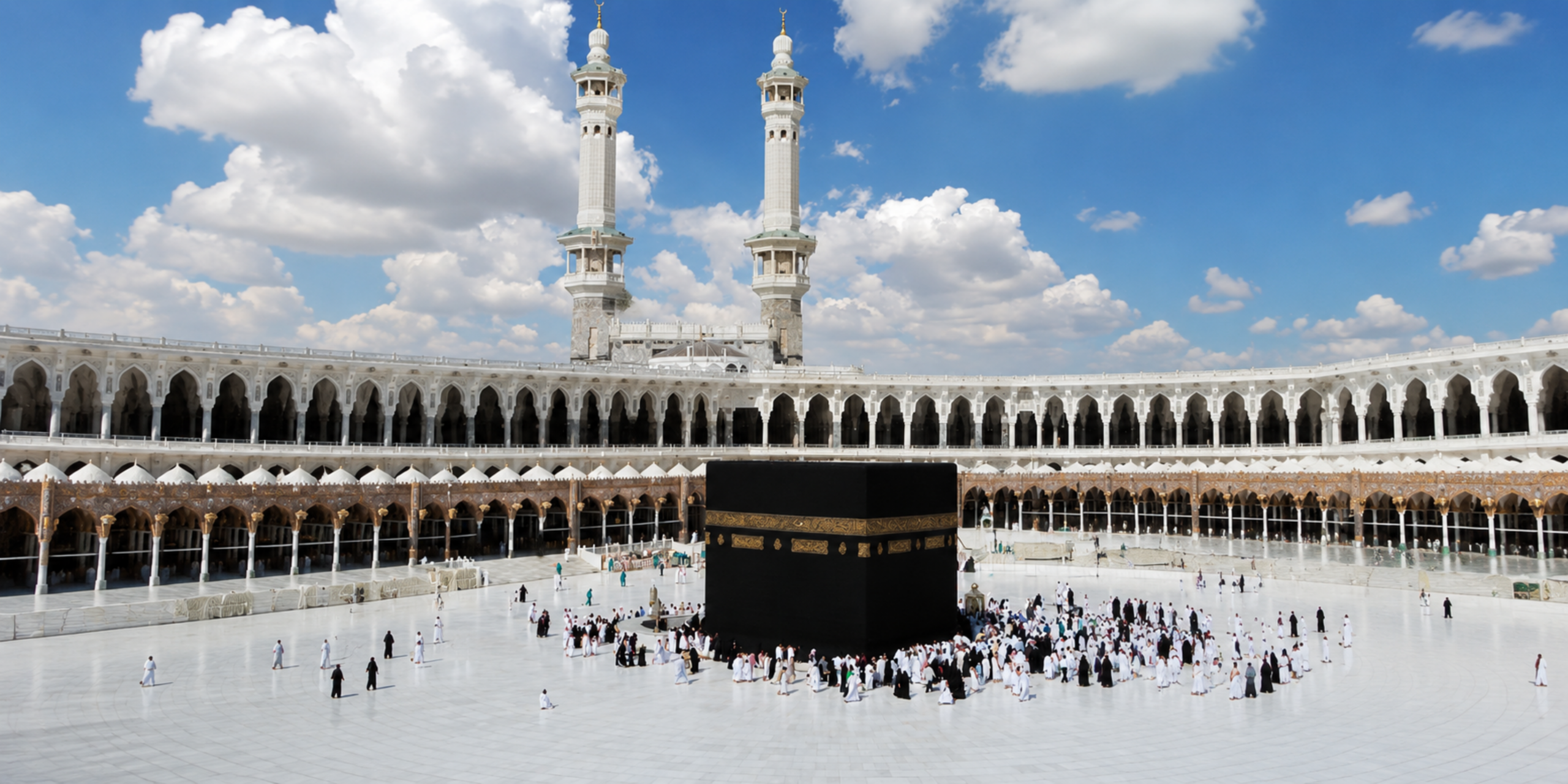 Une vue dégagée du Mataf montrant la Kaaba entourée de pèlerins sous un ciel bleu parsemé de nuages, encadrée par deux grands minarets et les arcades de la Grande Mosquée de La Mecque, illustrant la préparation et les précautions à prendre pour accomplir ses rites en été pour la Omra d'août 2027.