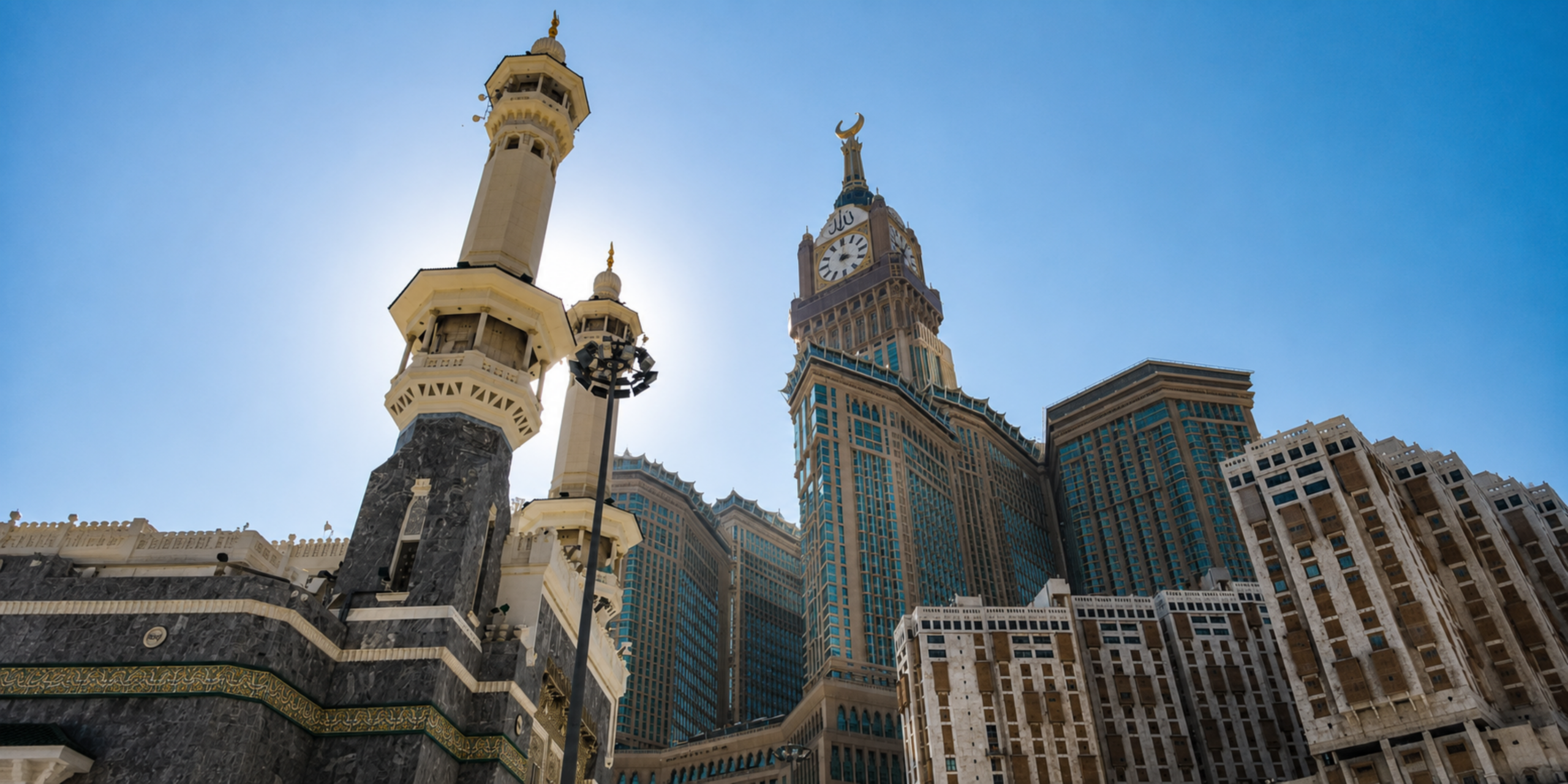 Une vue en contre-plongée de l'esplanade extérieure de la Grande Mosquée de La Mecque avec un minaret au premier plan, dominée par la Tour de l'Horloge et les complexes hôteliers environnants sous un ciel bleu, illustrant l'ensemble des services de transports, vols et hébergements inclus pour la Omra d'août 2027.