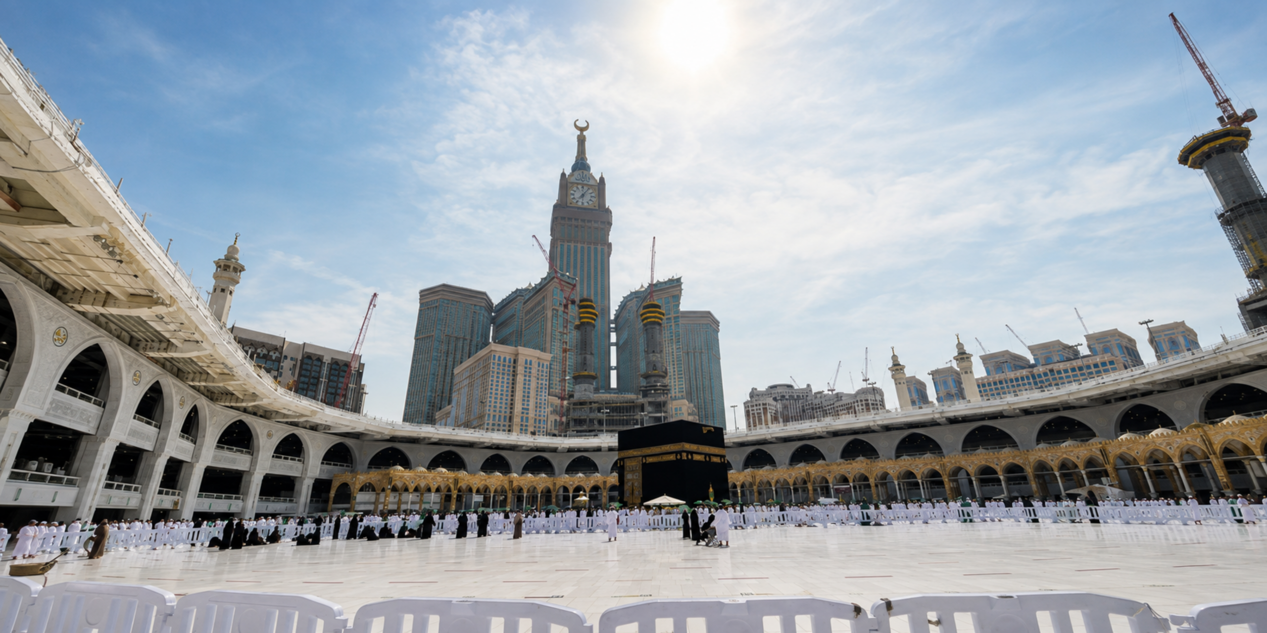 Une vue grand angle en contre-plongée de l'esplanade du Mataf baignée de soleil à La Mecque, montrant la Kaaba au centre entourée de barrières, les arcades de la Grande Mosquée et la Tour de l'Horloge s'élevant en arrière-plan sous un ciel voilé, illustrant l'organisation et les conseils pratiques pour effectuer sa Omra pendant le jeûne du Ramadan 2027.