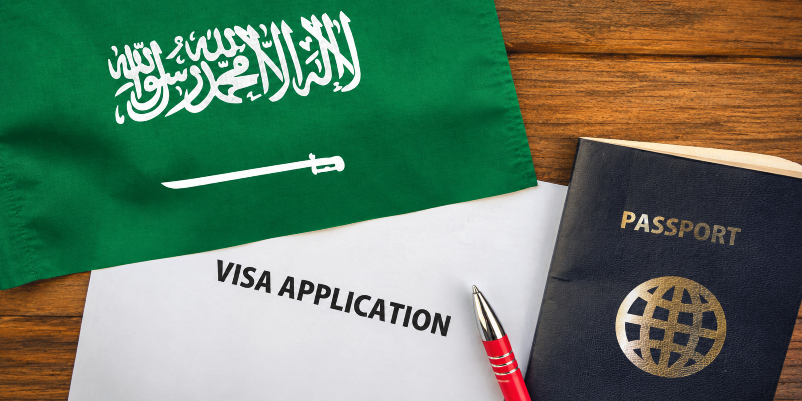Vue rapprochée sur une table en bois présentant un passeport, un formulaire de demande de visa avec un stylo rouge et le drapeau vert de l'Arabie Saoudite, illustrant les démarches administratives et la préparation des papiers pour la Omra du Ramadan 2027.