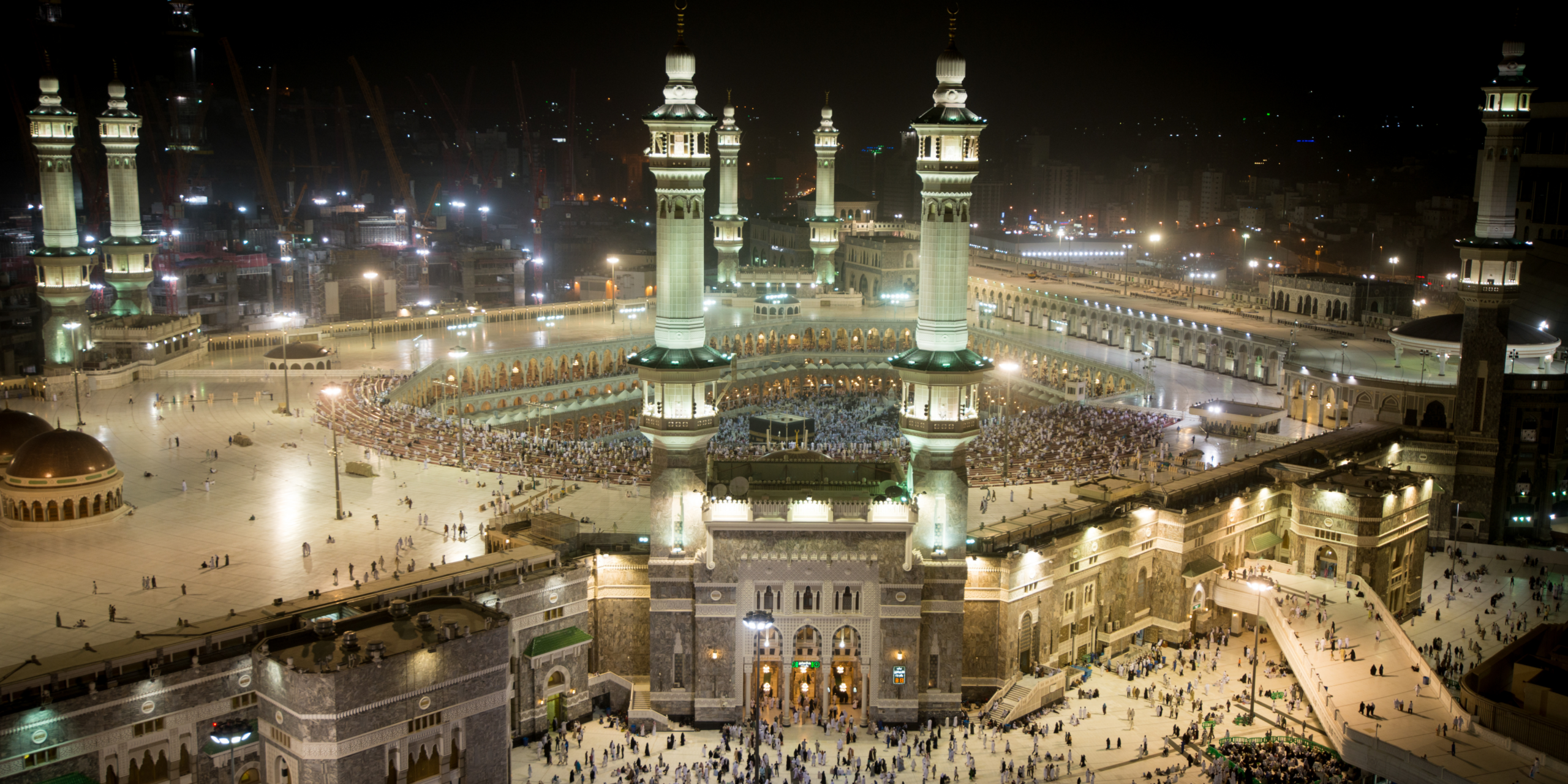 Une vue panoramique nocturne et majestueuse de la Mosquée Al-Haram à La Mecque, magnifiquement illuminée avec ses hauts minarets et le Mataf rempli de pèlerins au centre, illustrant la ferveur spirituelle et les mérites d'accomplir sa Omra pendant le mois béni de Ramadan 2027.