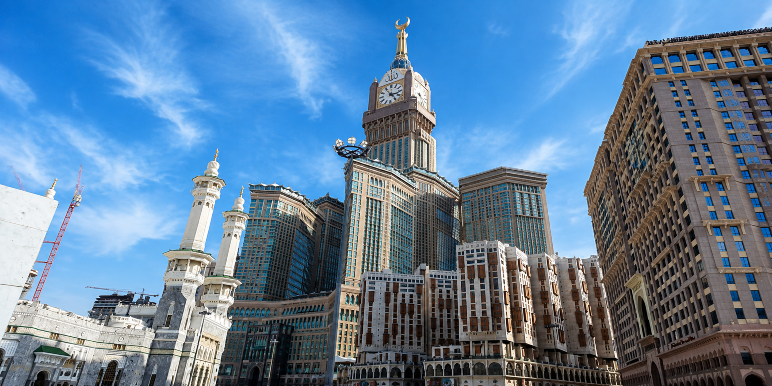 Une vue en contre-plongée des imposants complexes hôteliers et de la Tour de l'Horloge dominant l'esplanade extérieure et les minarets de la Grande Mosquée de La Mecque sous un ciel bleu ensoleillé, illustrant la gamme de services à la carte comprenant le logement, les transports, les visas et l'accompagnement pour la Omra du Ramadan 2027.