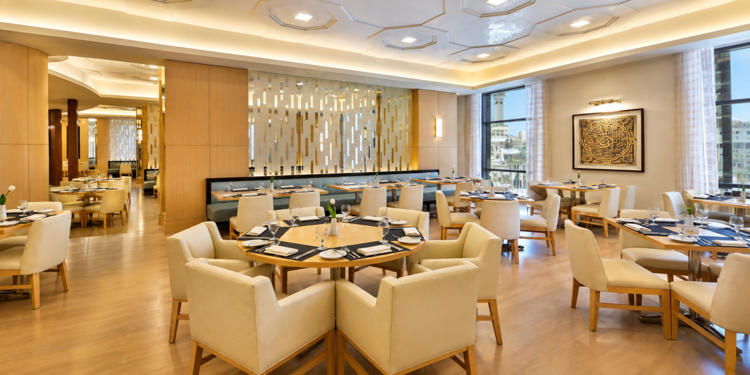 Une vue sur la salle à manger spacieuse et lumineuse du restaurant Al Mearaj, dotée de tables rondes et carrées soigneusement dressées, de sièges en cuir crème et de baies vitrées, illustrant l'offre gastronomique et les buffets raffinés de l'Hôtel Conrad Jabal Omar Makkah.