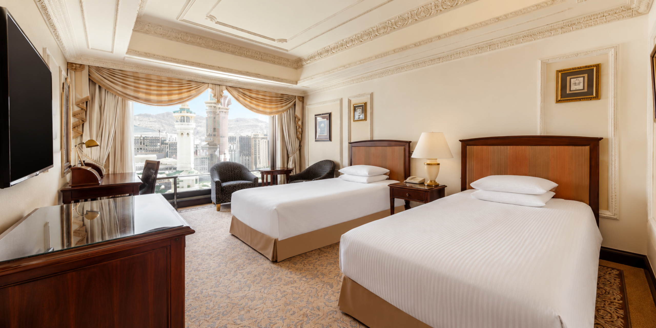Une chambre twin élégante comprenant deux lits simples préparés, un mobilier en bois massif et une large fenêtre offrant une vue directe sur les minarets du Masjid Al-Haram à La Mecque, illustrant le séjour idéal pour la Omra et le Hajj.