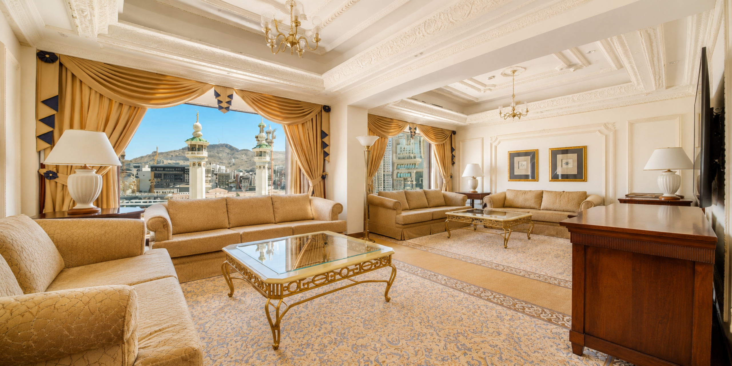 Un grand salon VIP luxueux décoré dans des tons dorés et beige, équipé de canapés confortables, de tables basses en verre et de grandes baies vitrées offrant une vue imprenable sur les minarets du Masjid Al-Haram à La Mecque, illustrant les installations de l'hôtel Dar Al Tawhid.