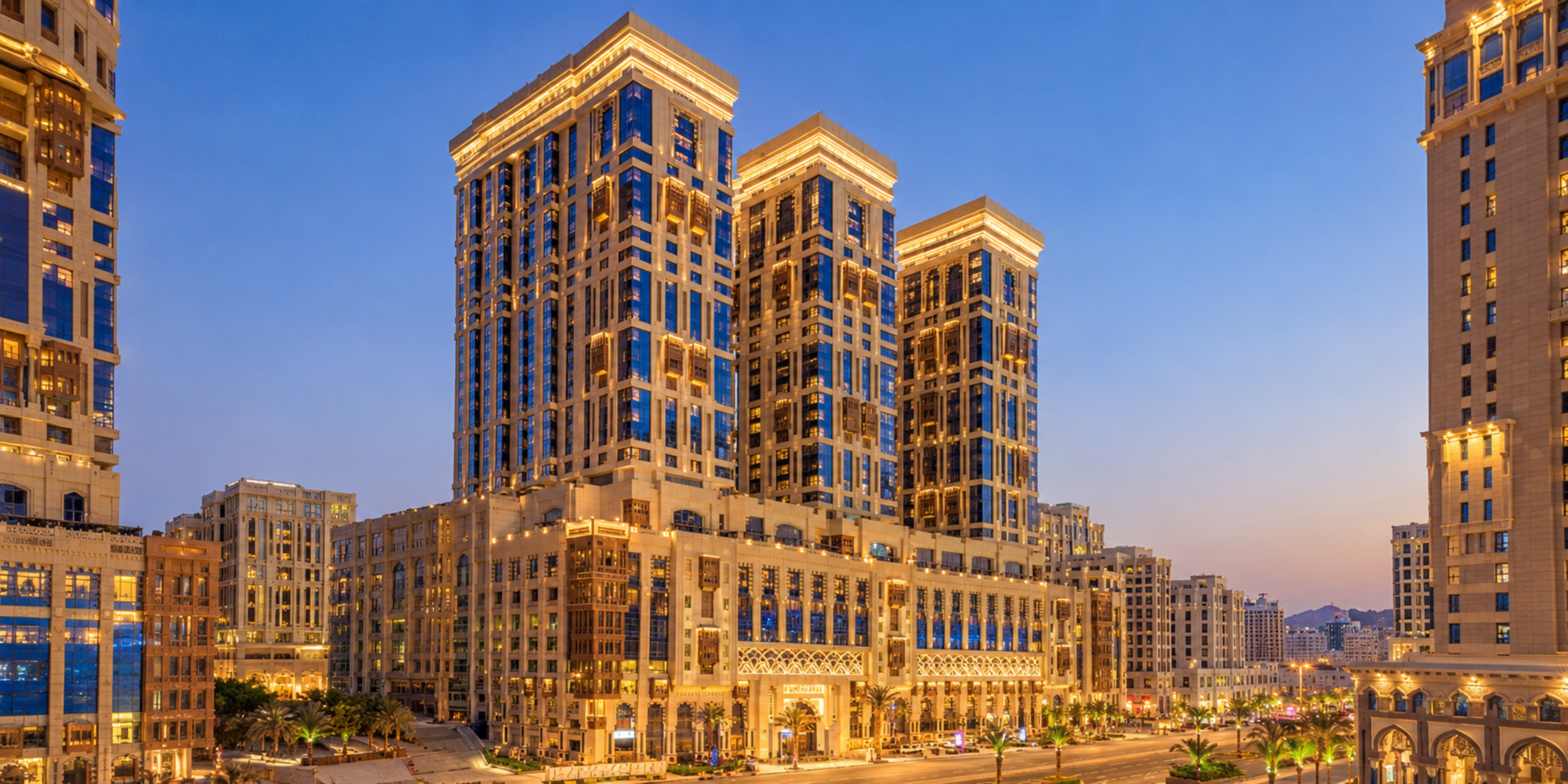 Vue extérieure des imposantes tours illuminées du Jabal Omar Hyatt Regency Makkah au crépuscule, illustrant la présentation et les atouts majeurs de cet hôtel de luxe idéal pour une Omra à La Mecque.
