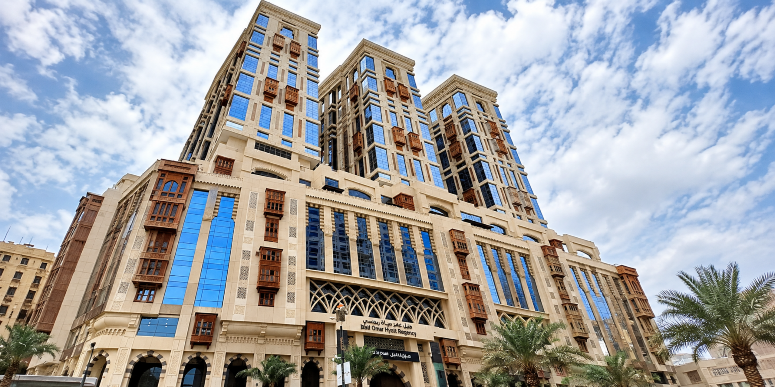 Vue en contre-plongée de la façade moderne de l'Hôtel Jabal Omar Hyatt Regency Makkah, illustrant son emplacement stratégique et son accès rapide au Masjid al-Haram pour les pèlerins.